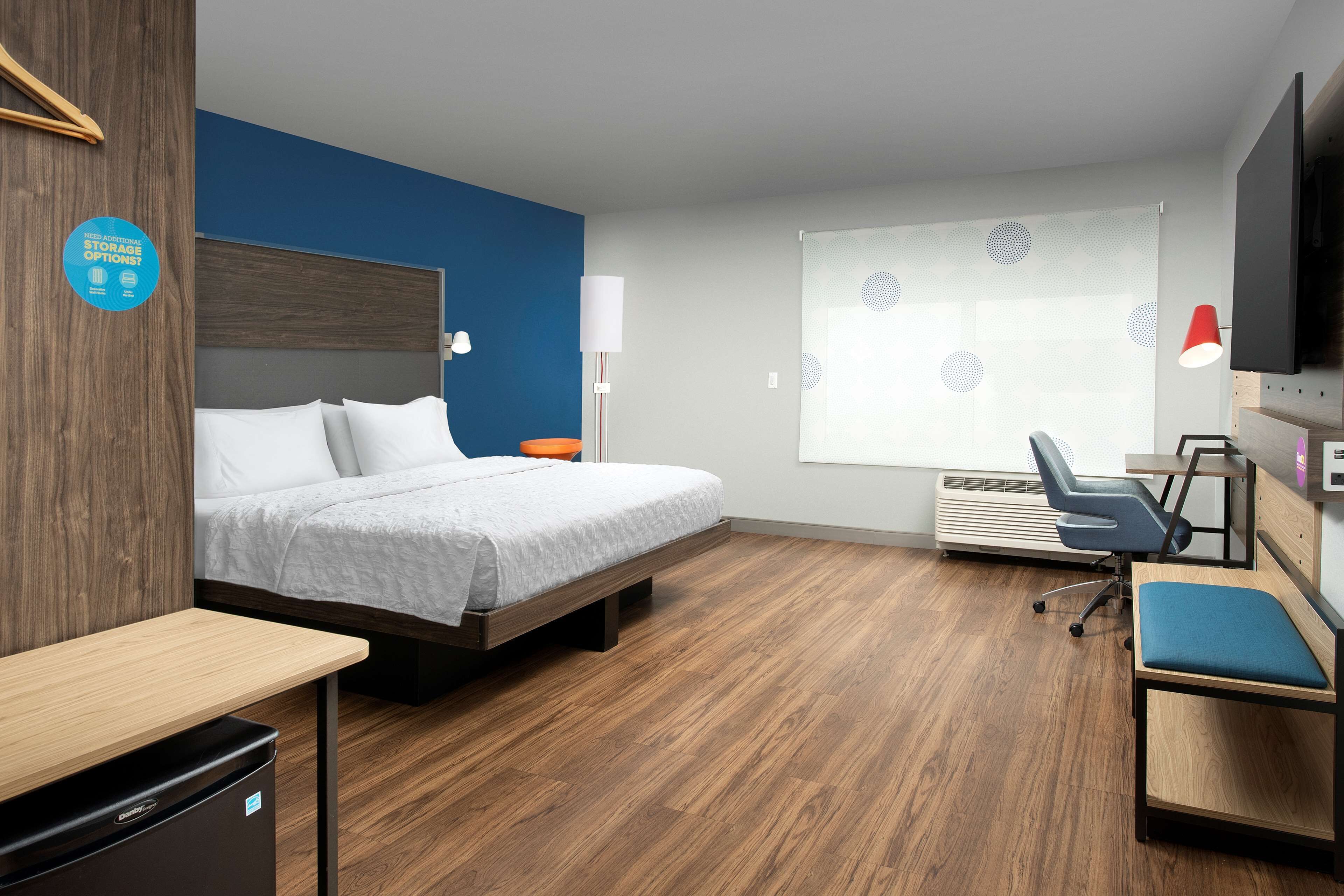 A modern hotel room with a bed, desk, chair, and a large window with blinds.