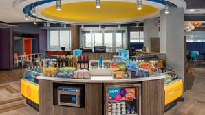 A hotel reception area with a counter displaying various beverages and snacks, a digital scale, and a 'Cool It' refrigerator. - Tru by Hilton Rapid City Rushmore (Rapid City)