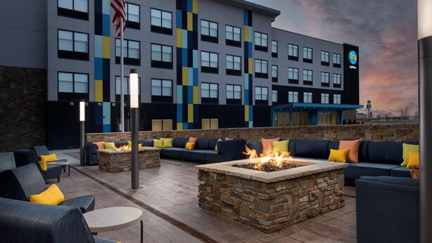 An outdoor seating area with sofas and round tables, a stone fire pit, and a building with a blue and yellow facade in the background. - Tru by Hilton Rapid City Rushmore (Rapid City)