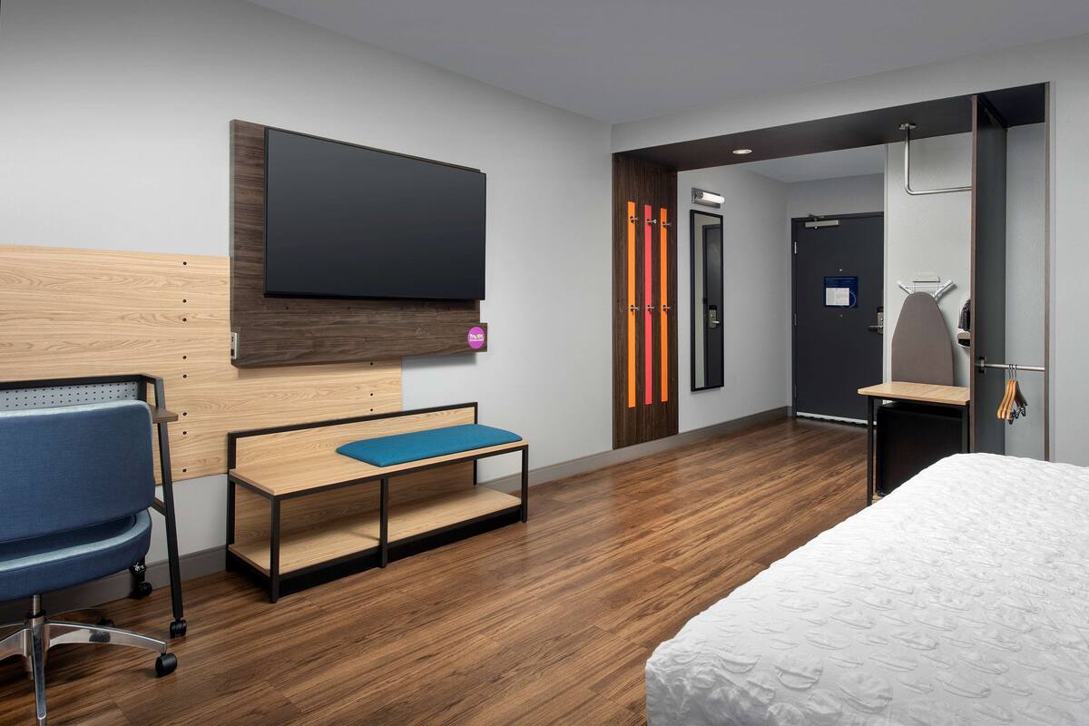 Une chambre d’hôtel moderne équipée d’un téléviseur à écran plat fixé au mur, d’un bureau en bois avec une chaise bleue, d’un lit avec une couette blanche et d’une porte donnant sur une salle de bain avec un porte-serviettes.