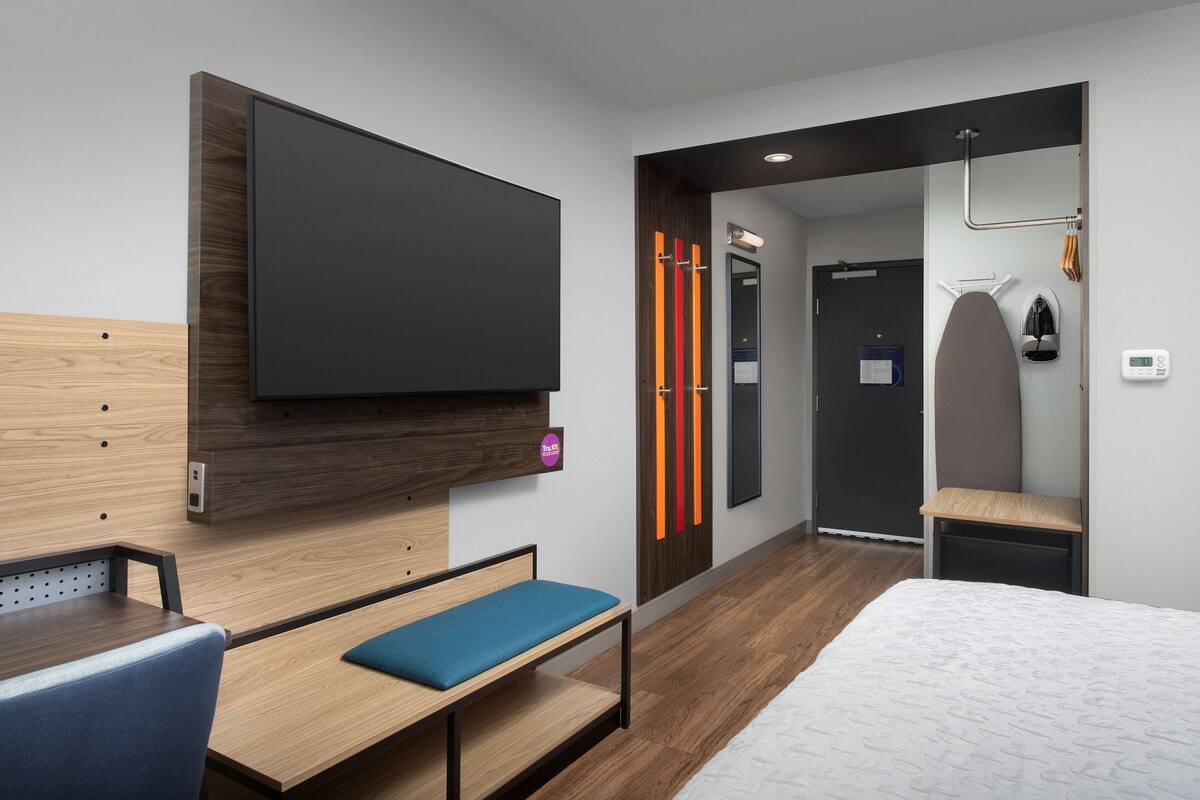 Une chambre d’hôtel moderne avec un téléviseur à écran plat fixé au mur, un banc en bois avec un coussin bleu, un lit recouvert d’une courtepointe blanche à motifs et une petite table avec une chaise bleue.
