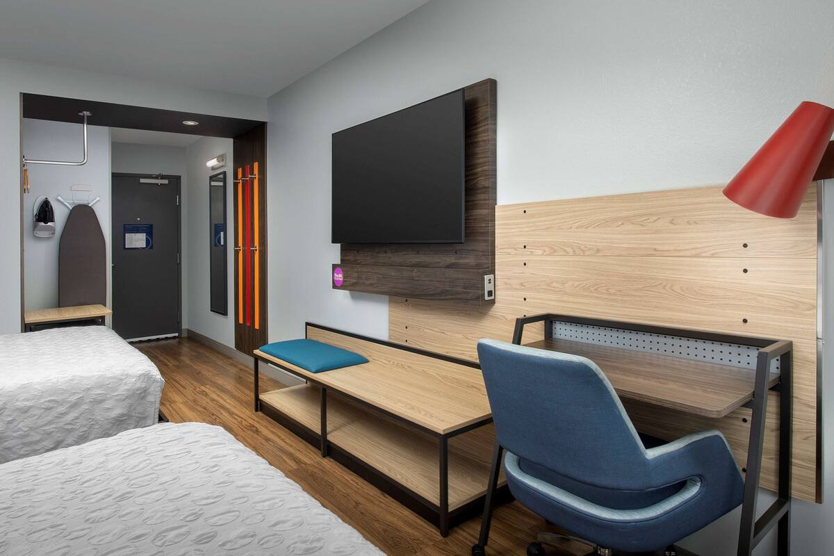 Une chambre d’hôtel moderne avec un bureau en bois, une chaise bleue, un téléviseur à écran plat et une lampe rouge.