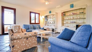 Un salon comprenant un canapé, un fauteuil et une table basse. On y trouve une bibliothèque avec des objets décoratifs et une fenêtre avec des rideaux.