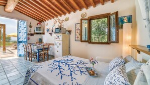 Un dormitorio con cama, zona de comedor con sillas y mesa, cocina con microondas y refrigerador, y ventana con vista a árboles.