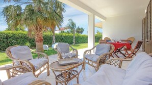 A covered outdoor seating area with wicker furniture and a table set for dining, overlooking a well-maintained garden and palm trees.