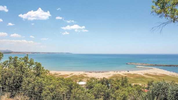 A coastal view with a beach, a pier, and a fenced area with trees.
