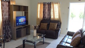 A living room with a glass coffee table, a brown leather sofa, a TV on a wooden stand, and a large window with curtains. - Your Holiday Home Caribbean Estates (Portmore)