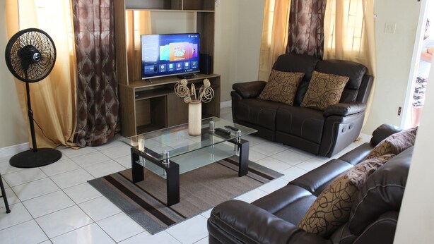 A living room with a sofa, a glass coffee table, a TV on a stand, a floor lamp, and a curtain. - Your Holiday Home Caribbean Estates (Portmore)