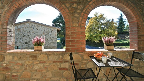 Un bâtiment en pierre doté de deux fenêtres en arc donnant sur un jardin. À l’intérieur, on trouve une table entourée de chaises et un saladier rempli de fruits.