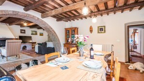 A dining area with a wooden table set for four, a brick fireplace, and a cozy living space with a TV and seating. - Lovely home in Suvereto -LI- (Suvereto -LI-)