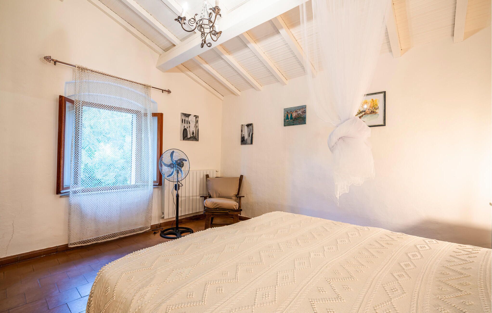 Ein Schlafzimmer mit einem großen Bett, einem Standventilator, einem Stuhl und einem Fenster mit durchsichtigem Vorhang.