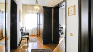 Un pasillo con suelo de madera, un candelabro, una silla, una alfombra y una puerta que da a otra habitación.