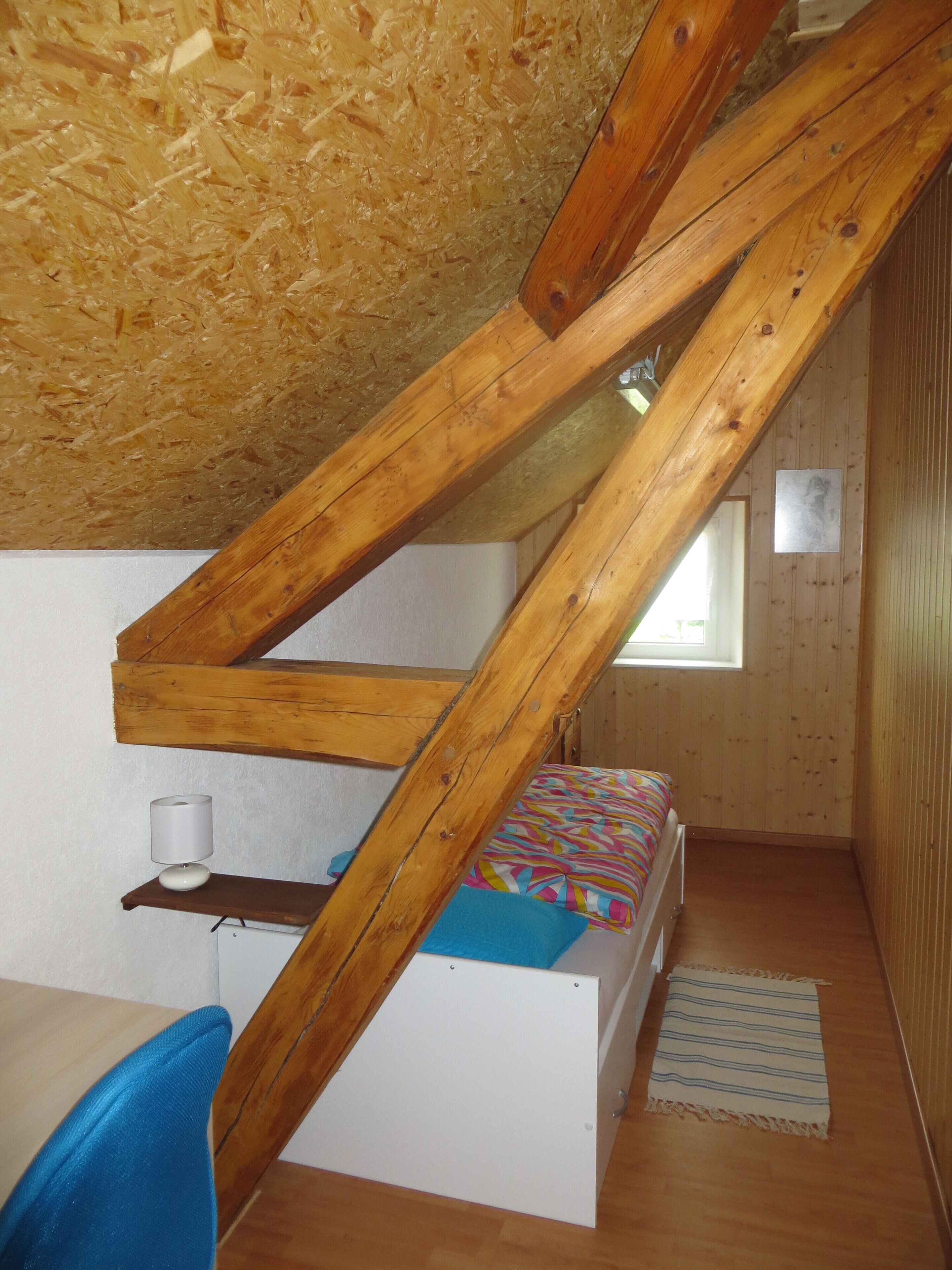 A small, wooden-paneled room with a sloped ceiling, a bed with colorful bedding, a small table with a lamp, and a window.