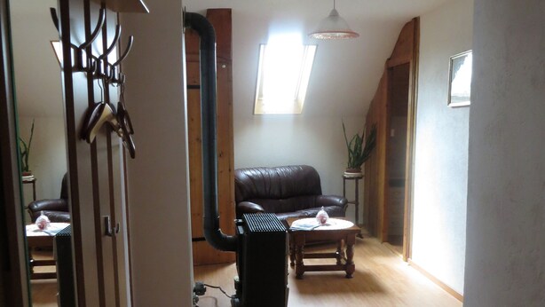Uma sala de estar ampla com teto alto, sofá de couro preto, mesa de centro de madeira e uma pequena mesa lateral. Há uma janela que proporciona iluminação natural e um ventilador de teto é visível.