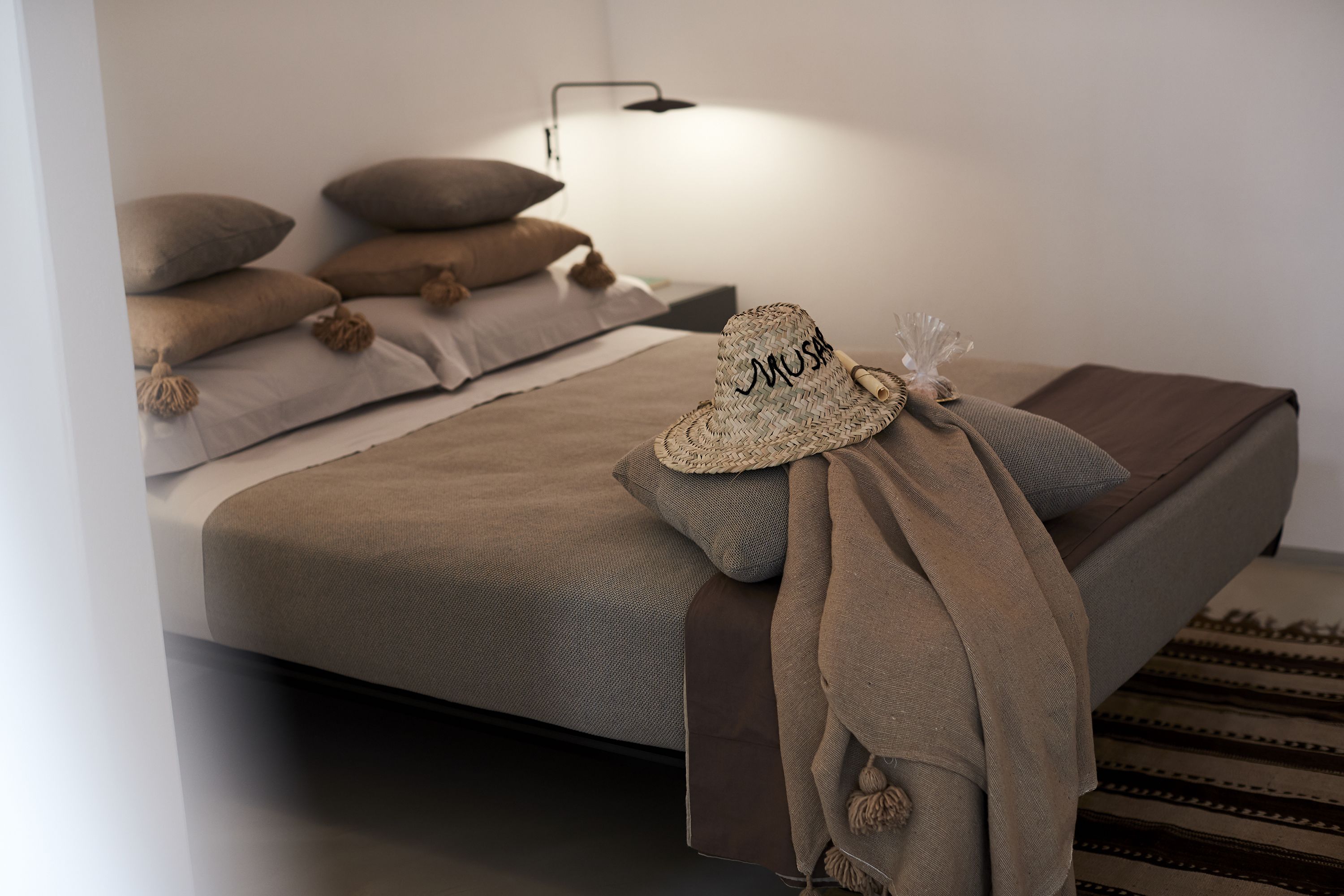 Una cama bien hecha con una manta color crema y un sombrero de paja con la palabra "MUSA".