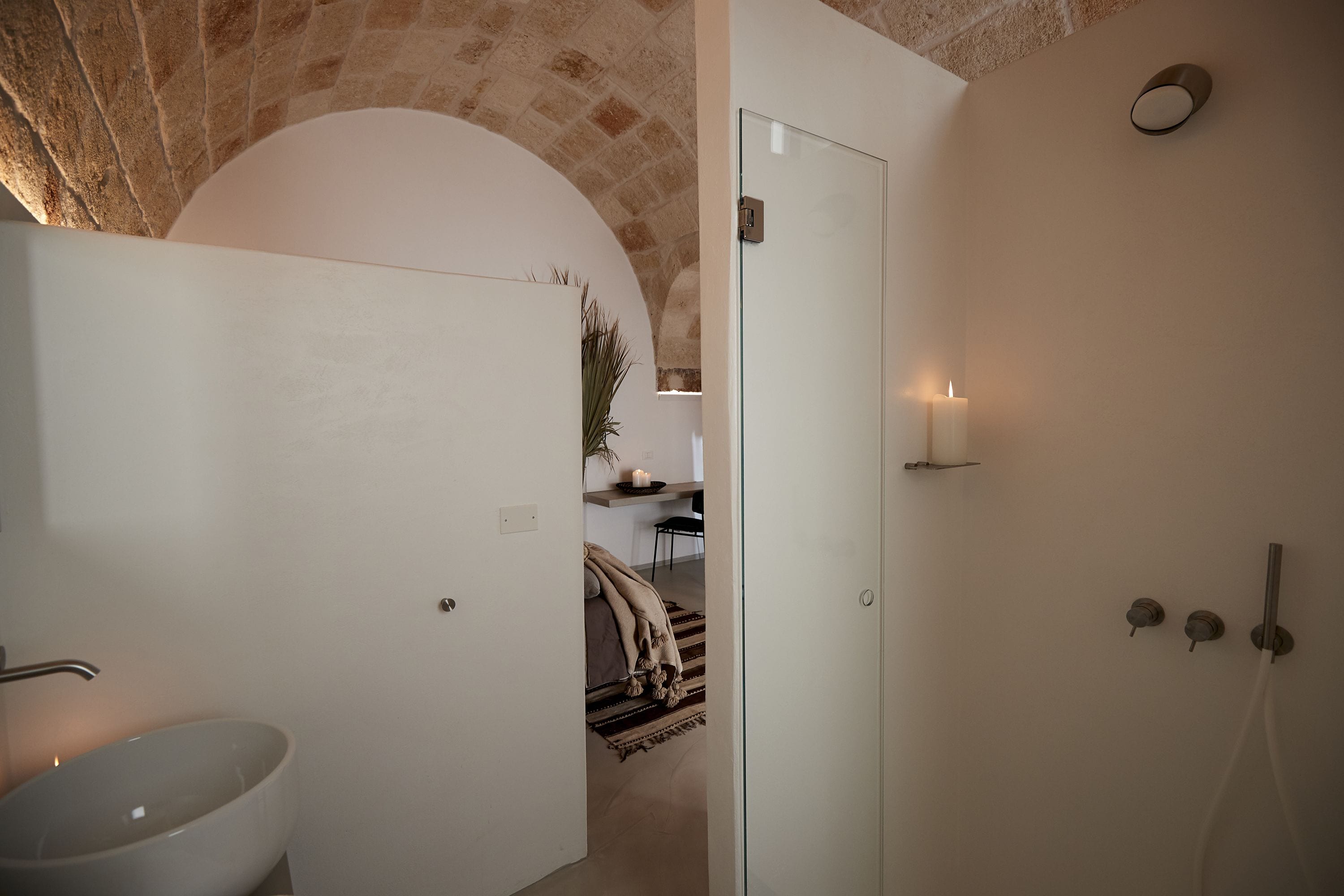 A bathroom with a white sink, a glass shower enclosure, and a door leading to a bedroom with a bed and a rug.