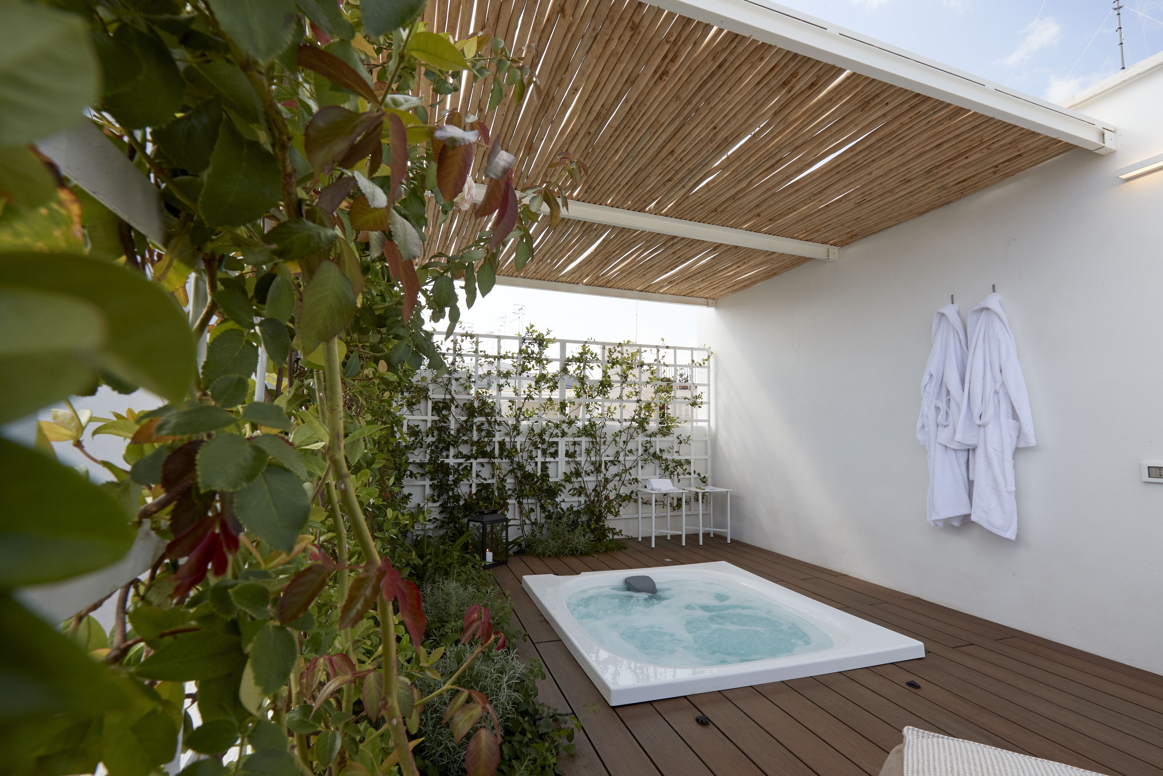 Een wellnessruimte met een jacuzzi, houten terras, witte muren en een witte omheining met beplanting.