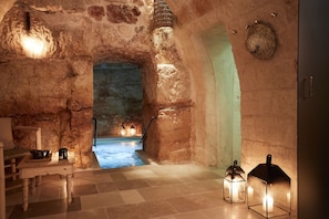 Una habitación con paredes de piedra, una piscina, una mesa de madera con cuencos y dos faroles.
