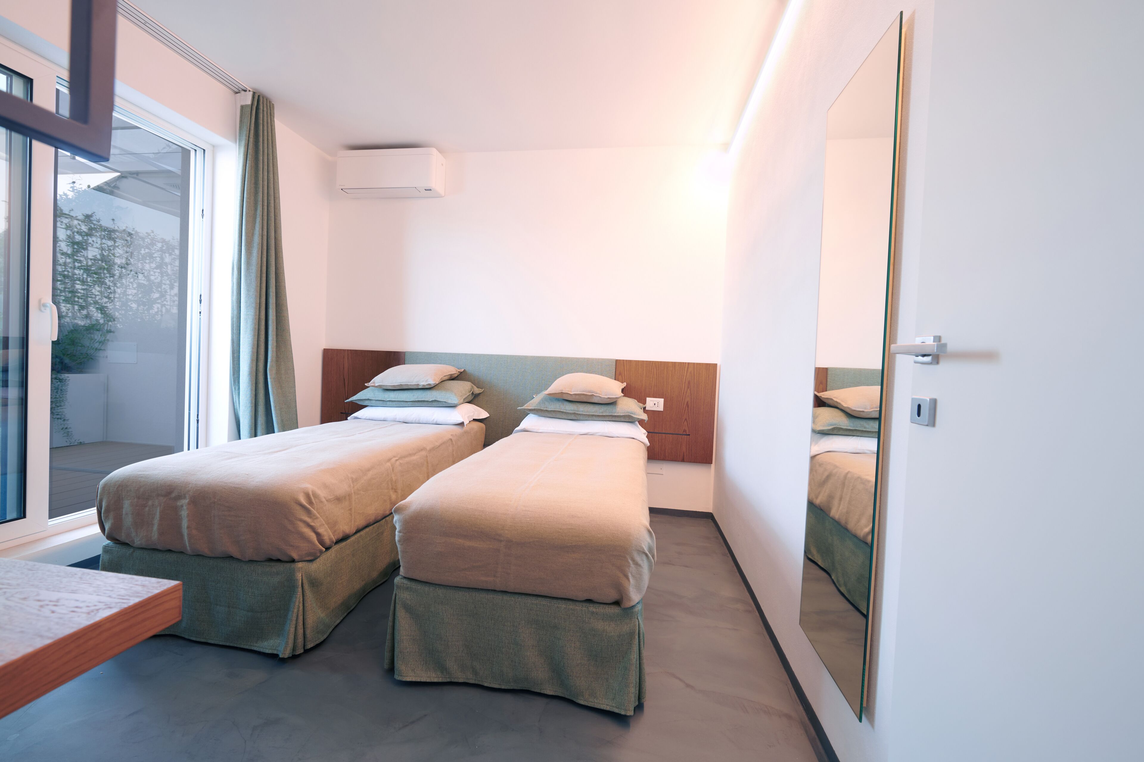 A modern hotel room with two single beds, a large mirror, and a sliding glass door to the outside.