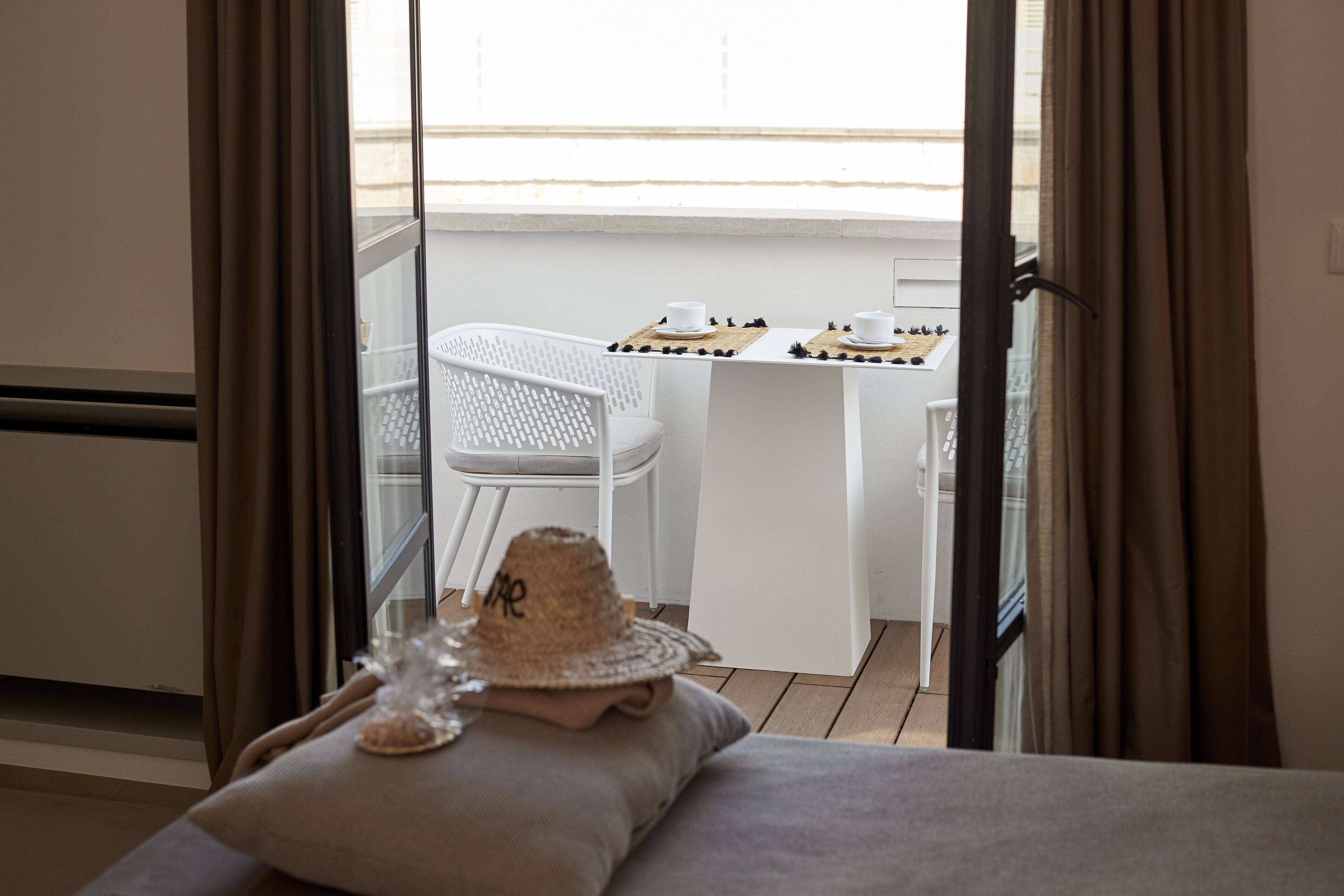 A balcony with a white chair, a small table with cups and a hat, and a view of the outside.