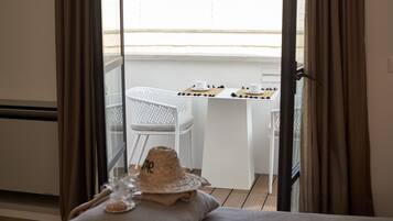 A balcony with a white chair, a small table with cups and a hat, and a view of the outside.