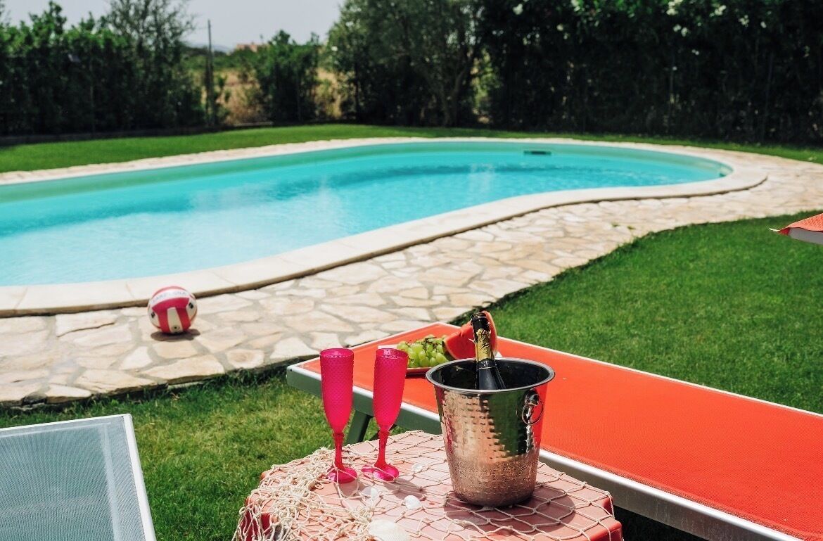 Une scène au bord d’une piscine, avec une eau claire, une table sur laquelle se trouve une bouteille et deux verres.