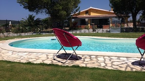 A swimming pool with two red lounge chairs, a stone-tiled deck, and a yellow building in the background. - Stunning House Between the Countryside and the Sea - Southern Sardinia CIN: IT111070C2000U0960 (Porto Pino (Sardegna))