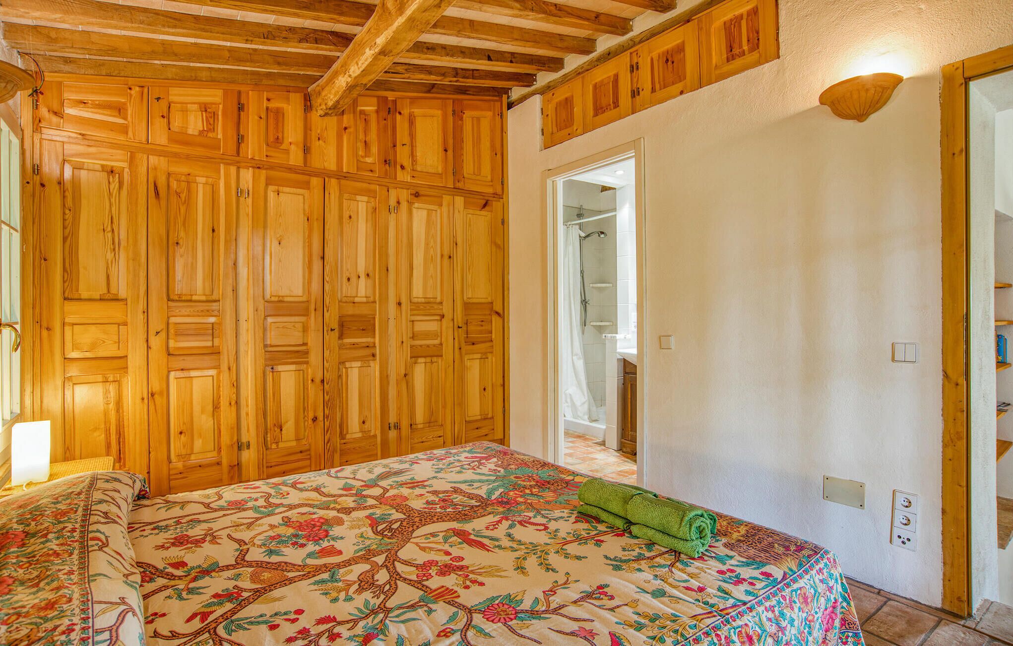 Une chambre avec des lambris en bois, un lit recouvert d’une courtepointe à motifs floraux et une porte de salle de bain visible.