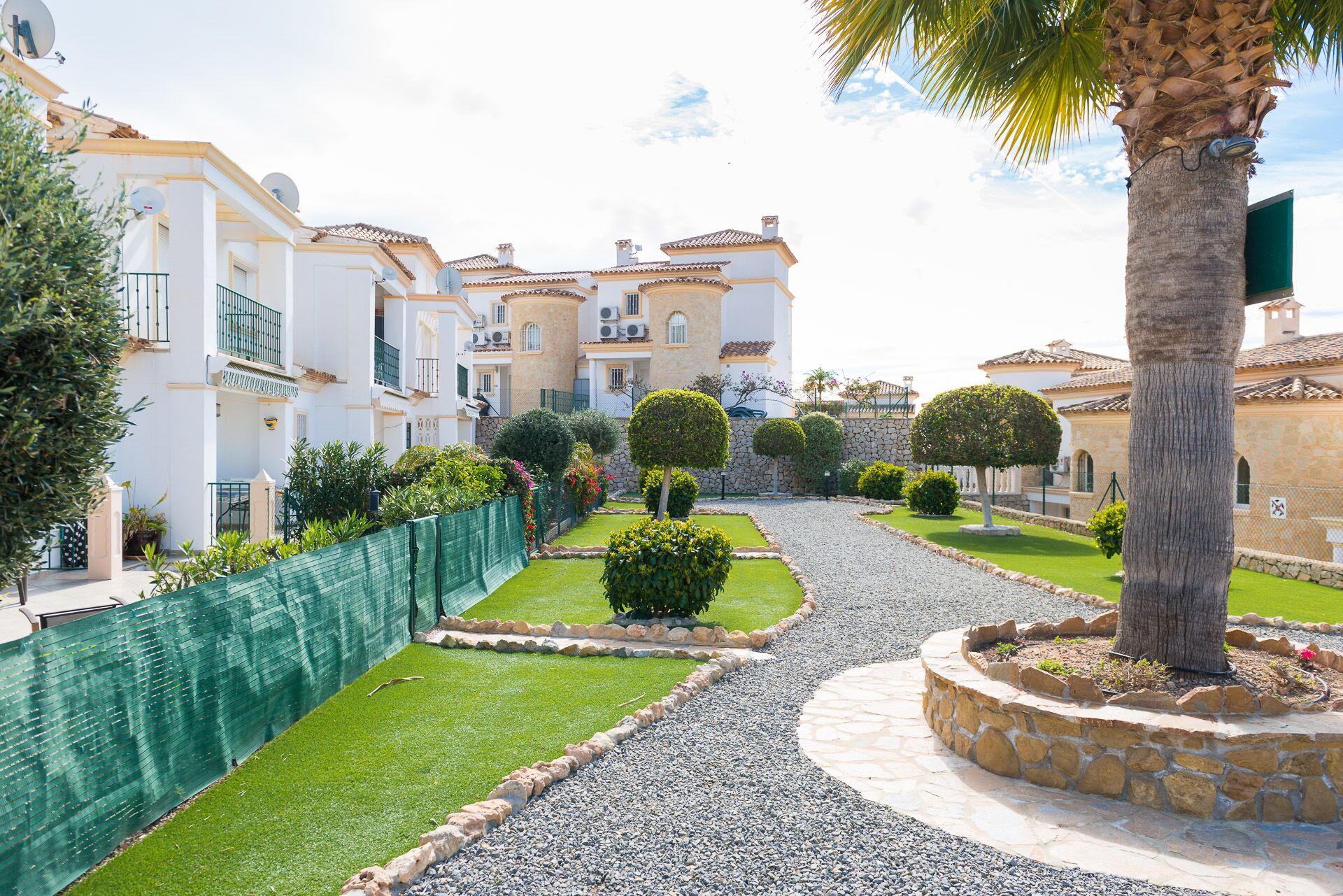 Un quartier résidentiel avec un chemin pavé, des haies bien taillées et un palmier.