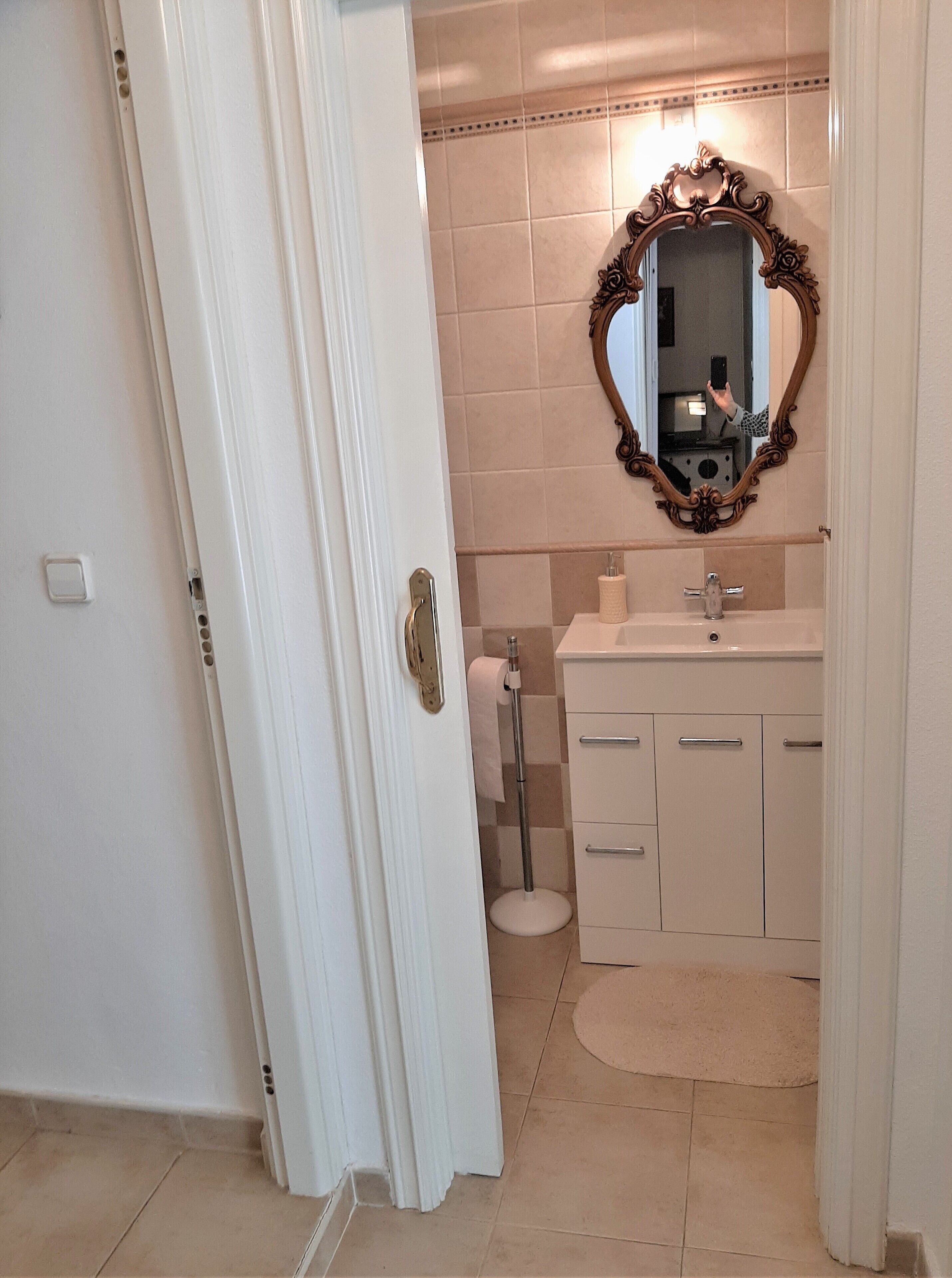 A bathroom with a white door, beige tiles, a mirror with an ornate frame, a sink, and a toilet.