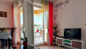 A modern living room with a glass coffee table, a flat-screen TV on a white cabinet, a dining area with a table and chairs, and a balcony with potted plants and a red chair. - Apartment near sea and city (Antibes)