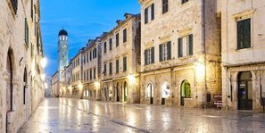 Une rue historique bordée d’immeubles en pierre, avec une tour imposante et un pavé mouillé qui reflète les lumières.