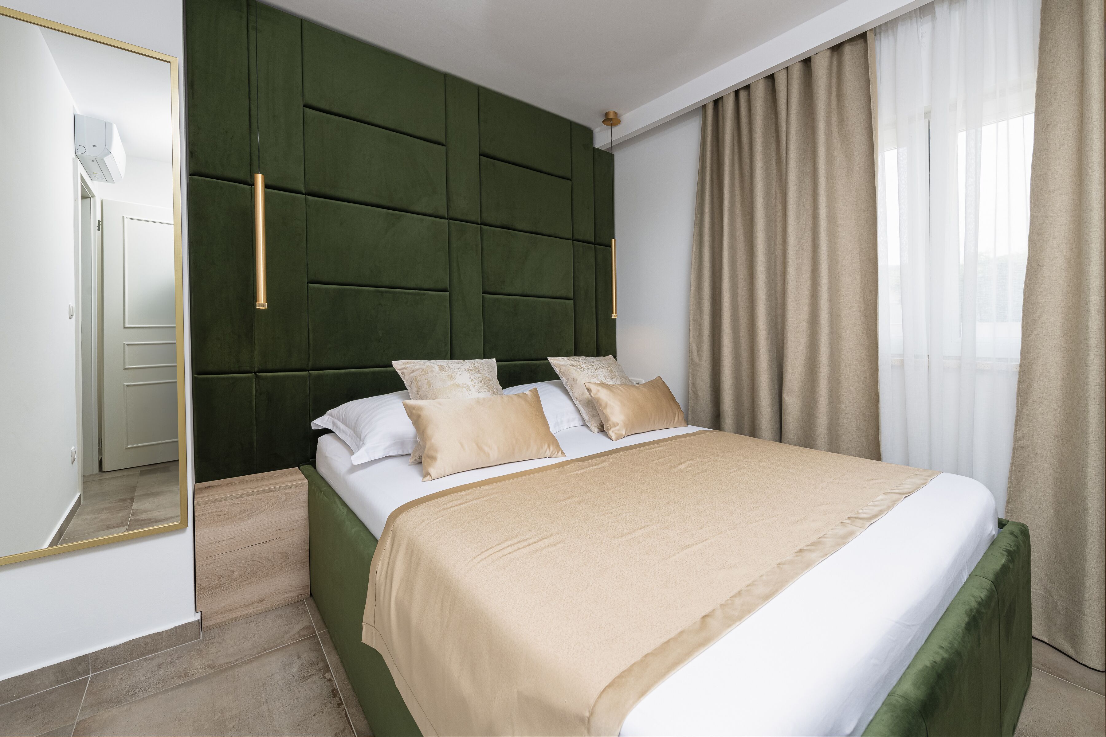 A neatly made bed with a green headboard, beige bedding, and a mirror reflecting part of the room.