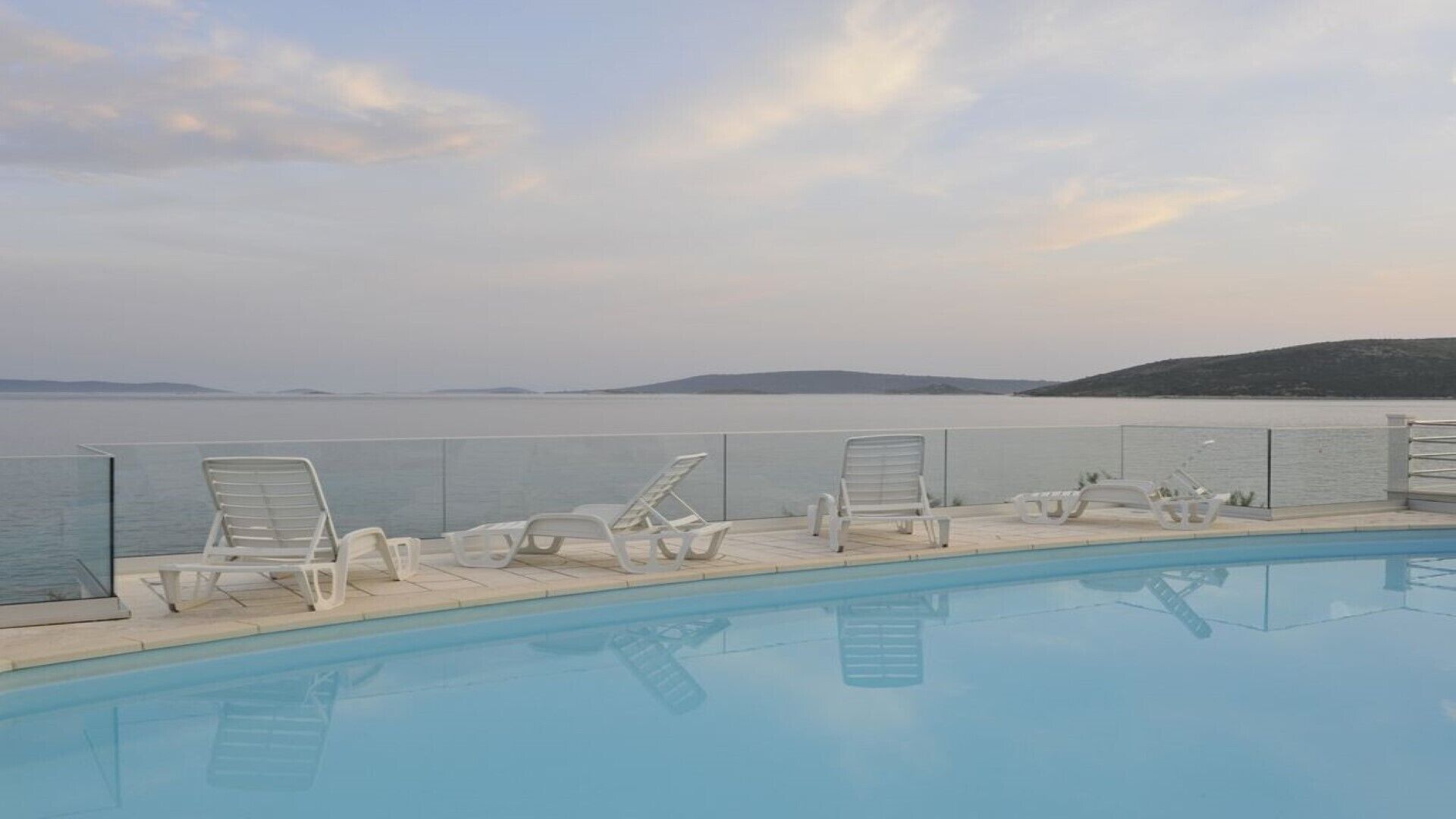 A pool with lounge chairs facing a calm sea and distant hills.