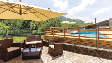 Patio con mobili in vimini e tavolo in vetro, con vista su un'area piscina con recinzione in legno e ombrelloni.