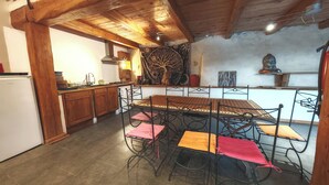 A rustic kitchen with wooden beams, a dining table with metal chairs, and a decorative tapestry. - SMALL GITE "BERGERIE" (Bargeme)