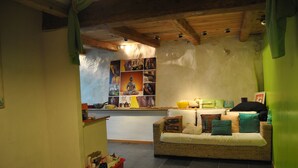 A cozy living area with a wicker sofa, a small kitchen area in the background, and a red storage bin on the floor. - SMALL GITE "BERGERIE" (Bargeme)