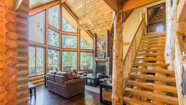 A log cabin interior with a fireplace, wooden staircase, and large windows.