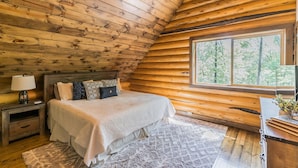 A cozy bedroom with a wooden ceiling, a large window, a bed with a white comforter, and a nightstand with a lamp.