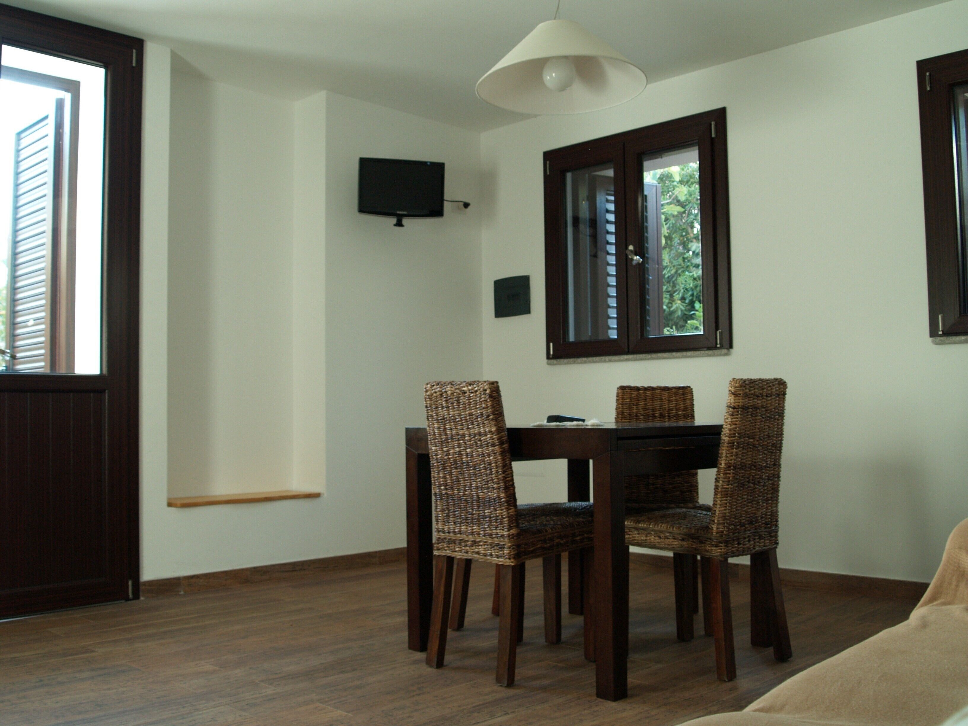 A dining area with a wooden table and four wicker chairs, a television mounted on the wall, and a window with a view of greenery.