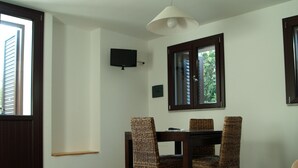 A dining area with a wooden table and four wicker chairs, a television mounted on the wall, and a window with a view of greenery.