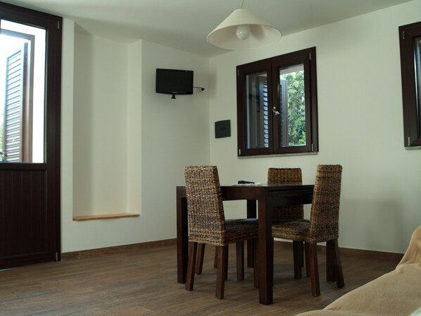 A dining area with a wooden table and four wicker chairs, a television mounted on the wall, and a window with a view of greenery.