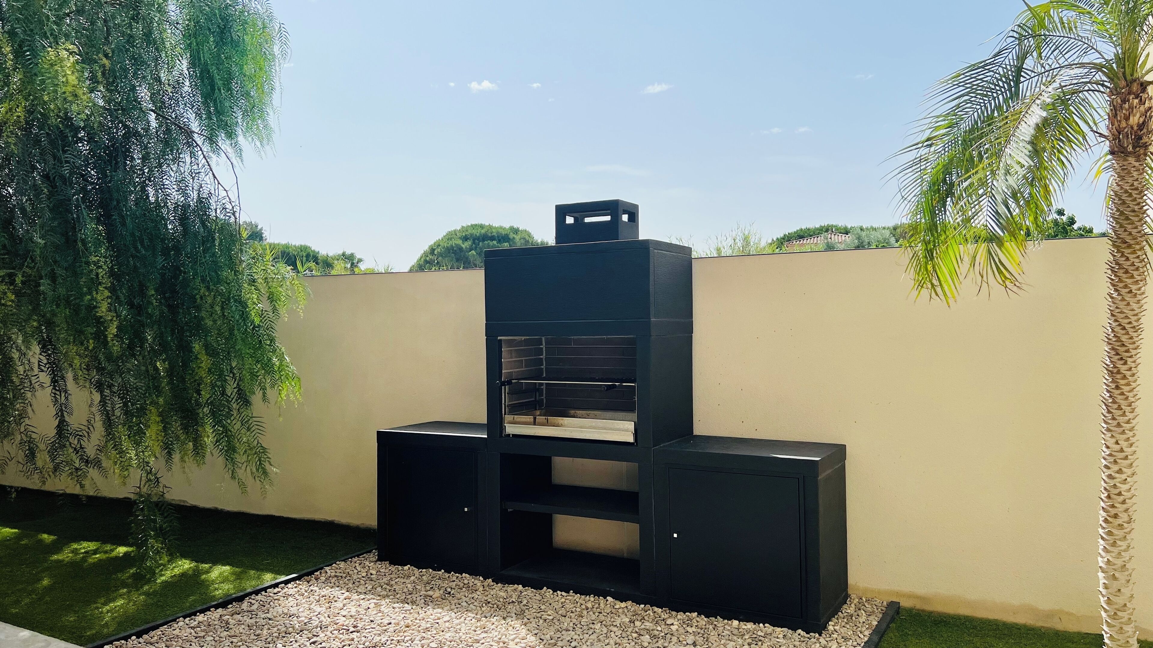 An outdoor barbecue grill with a chimney, surrounded by a low wall, gravel, and greenery.