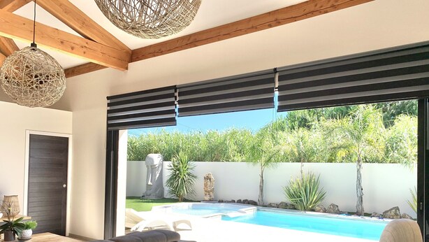 A modern living room with a grey sofa, a coffee table, and a view of a swimming pool and garden. - Pool Villa Near Beach (Agde)