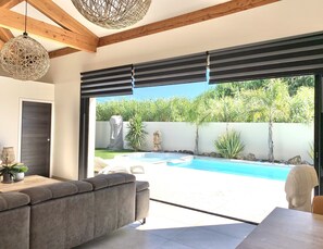 Een moderne woonkamer met een grijze bank, een salontafel en uitzicht op een zwembad en tuin.