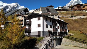 Un chalet de montagne doté d’un escalier en pierre menant à l’entrée, entouré de montagnes enneigées et surplombant un ciel bleu azur.