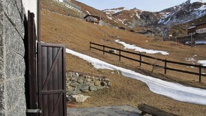 Une entrée dallée de pierres, avec un portail en bois, un mur de pierres et une vue sur un paysage montagneux parsemé de plaques de neige.