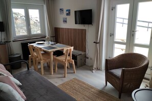A cozy room with a bed, a dining table with chairs, a wicker chair, a TV, and a window with curtains.