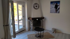 Une pièce avec une cheminée en pierre, une horloge murale ronde, un tableau encadré, un petit tabouret et une table ronde avec un pied en fil de fer.