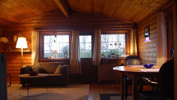 Ein gemütliches Blockhaus-Interieur mit Holzbalkendecke, einer Couch, einem Esstisch mit Stühlen und einem Fenster mit Vorhängen.